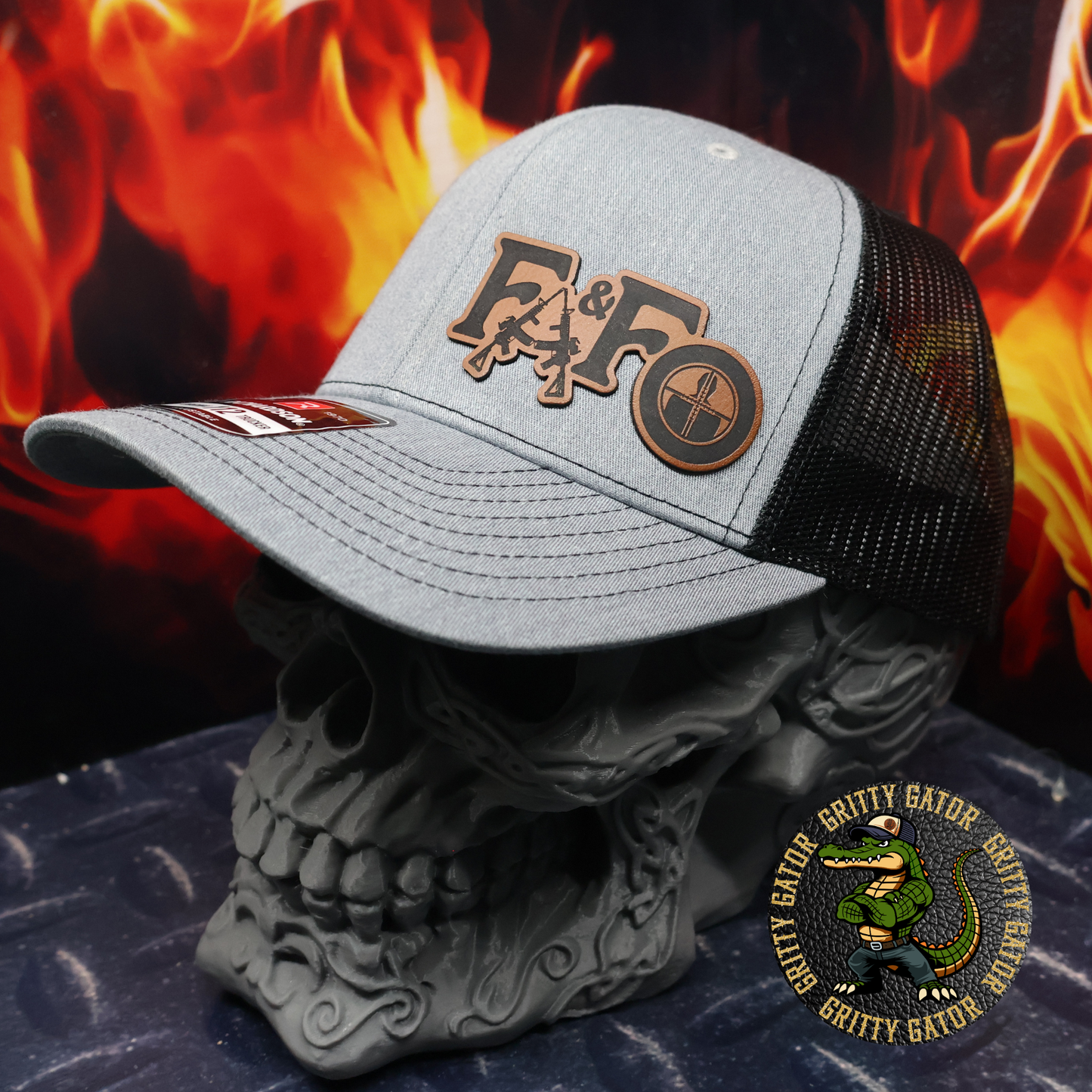 Heather gray and black Richardson 112 trucker hat with a leatherette patch featuring guns and a reticle reading “FAFO,” displayed on a skull stand with flames in the background, by Gritty Gator.