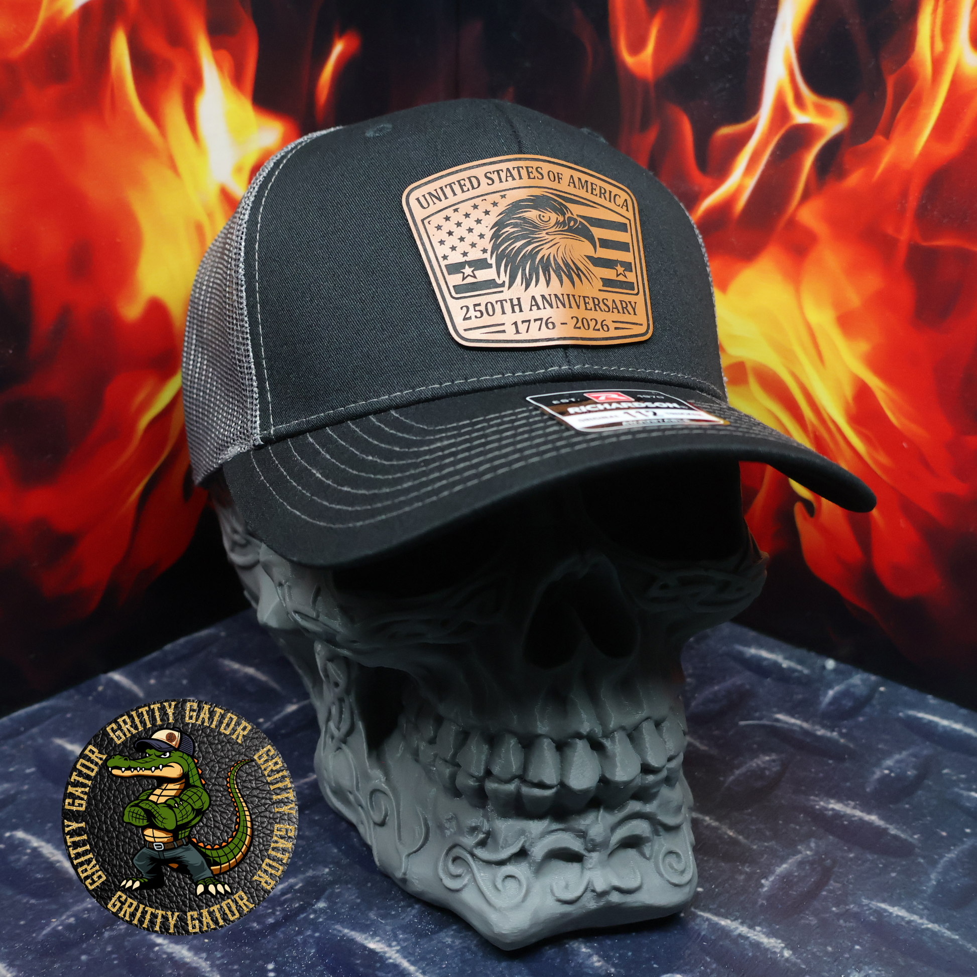 Black and charcoal Richardson 112 trucker hat with a leatherette patch featuring a bald eagle and American flag reading “United States of America – 250th Anniversary 1776–2026,” displayed on a skull stand with flames in the background, by Gritty Gator.