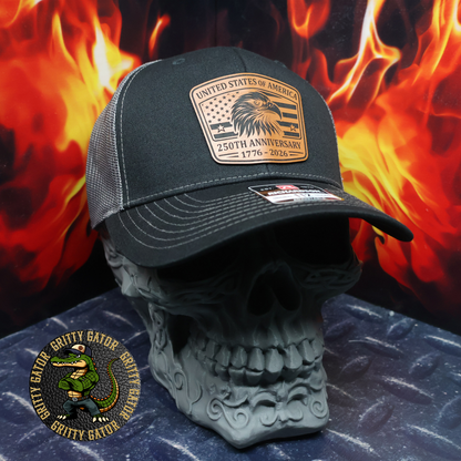 Black and charcoal Richardson 112 trucker hat with a leatherette patch featuring a bald eagle and American flag reading “United States of America – 250th Anniversary 1776–2026,” displayed on a skull stand with flames in the background, by Gritty Gator.
