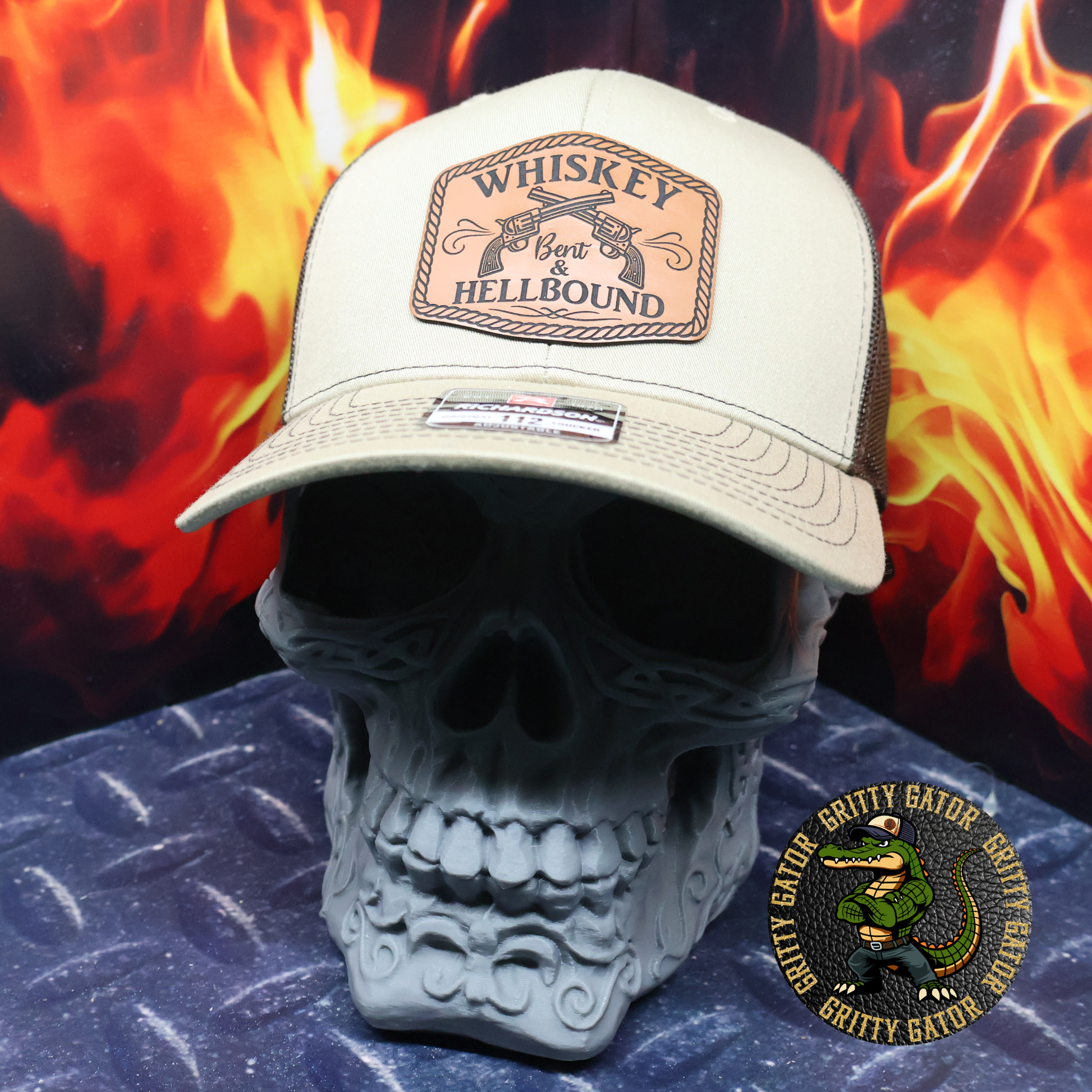 Khaki and coffee Richardson 112 trucker hat with a leatherette patch reading “Whiskey Bent & Hellbound” featuring crossed revolvers, displayed on a skull stand with flames in the background, by Gritty Gator.